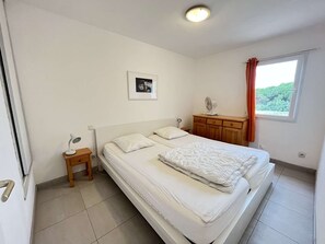 A bedroom with a bed, a nightstand, a small table, a fan, a window with curtains, and a picture on the wall.