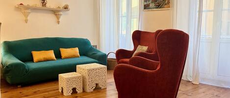 Un salon avec un canapé turquoise, un fauteuil rouge et deux petites tables d’appoint blanches ornées de motifs complexes.