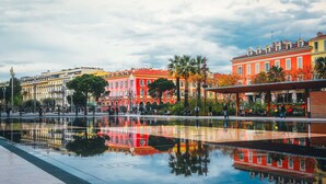 Un élément décoratif aquatique, conçu pour créer des reflets, dans une place publique, avec des bâtiments aux couleurs vives en arrière-plan.