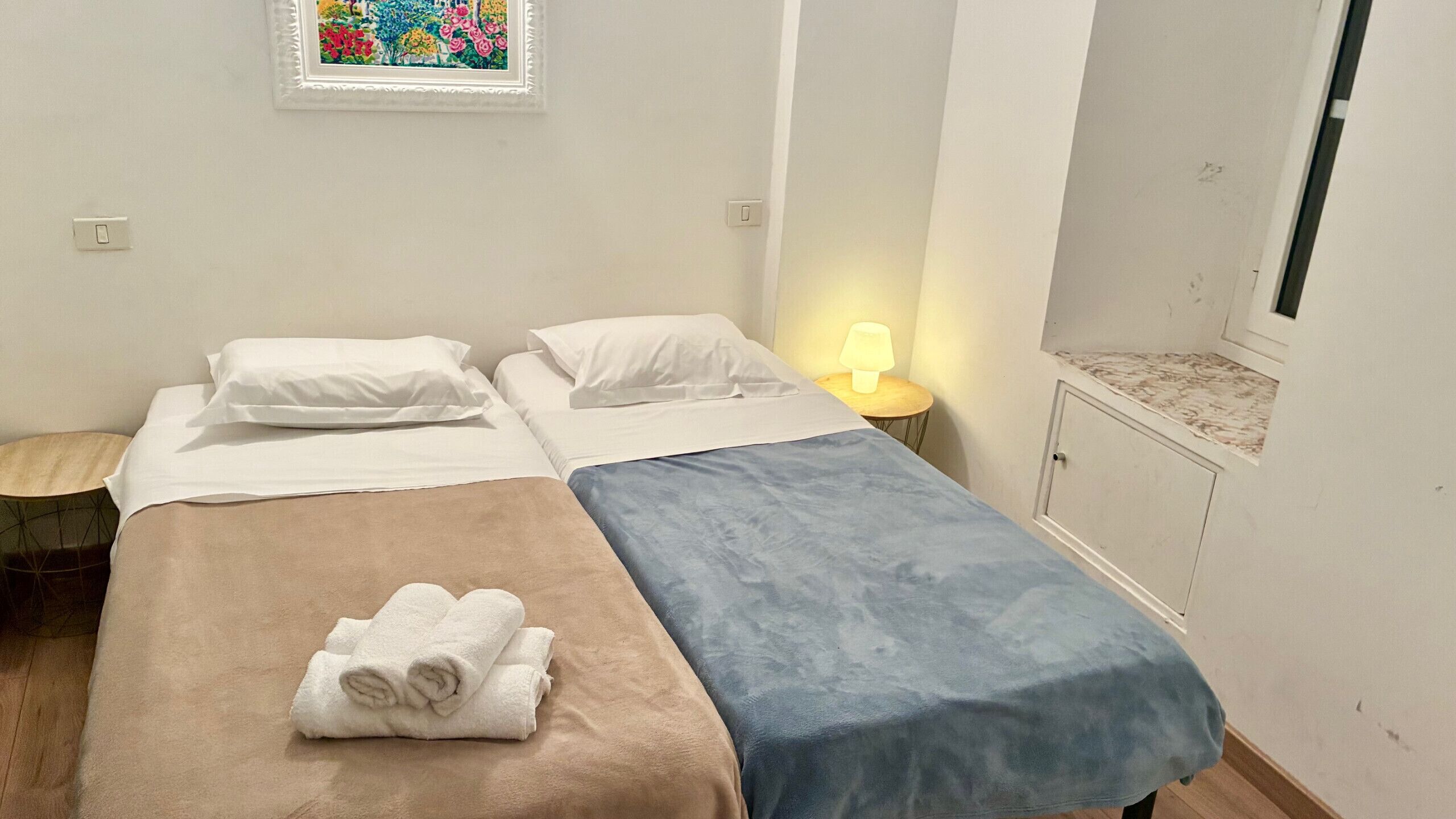 Une chambre double avec deux lits, une petite table avec une lampe, une armoire et un tableau coloré au mur.