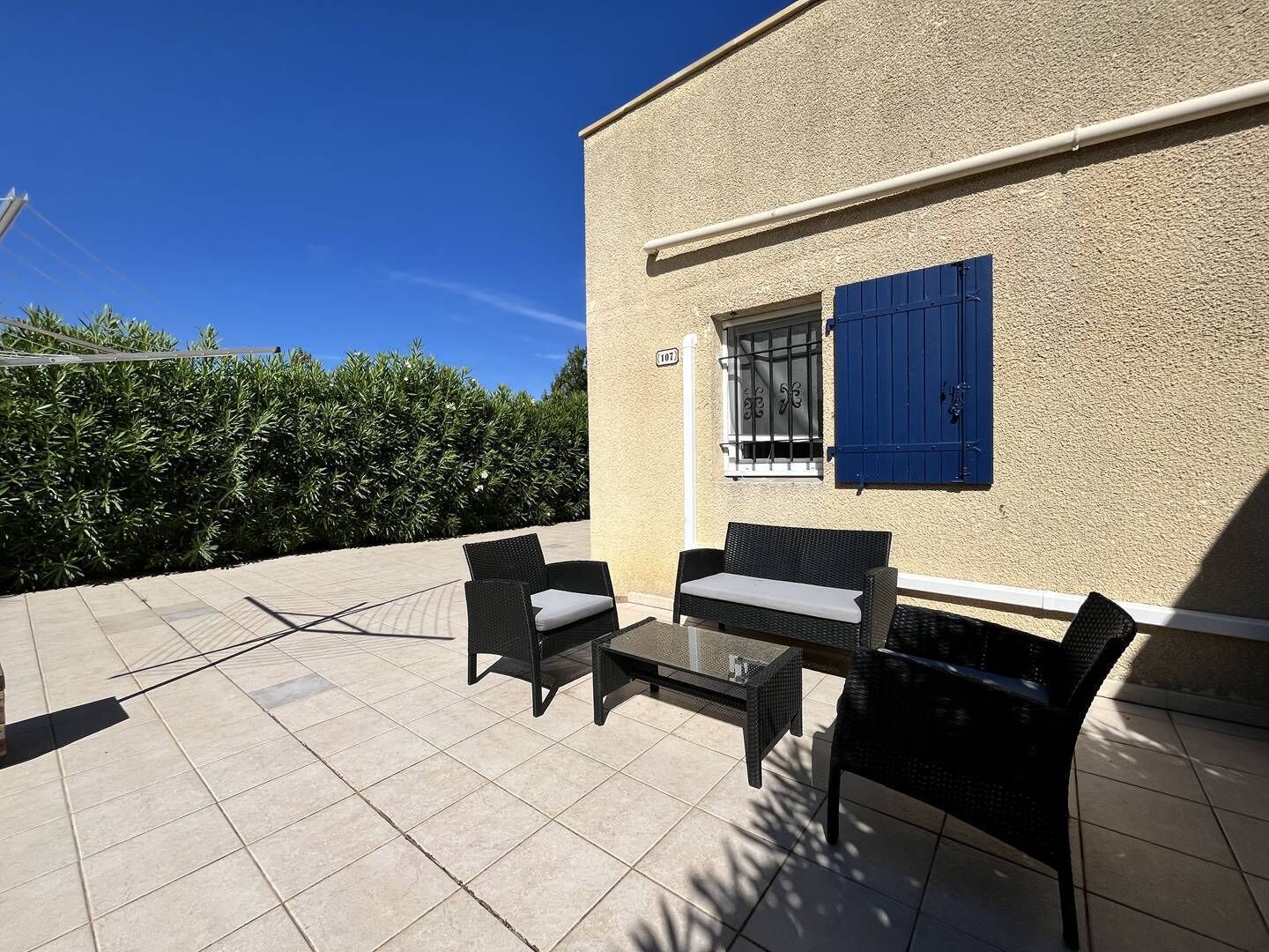 Une terrasse extérieure avec un sol carrelé, deux fauteuils en osier, une petite table et une fenêtre à volets bleus.