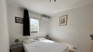Une chambre à coucher comprenant un lit, une table de chevet, une lampe, un appareil de climatisation et une fenêtre avec des rideaux.