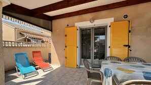 Eine überdachte Terrasse mit zwei Liegestühlen, einem Tisch mit Tischdecke und einer Glastür mit gelben Fensterläden.