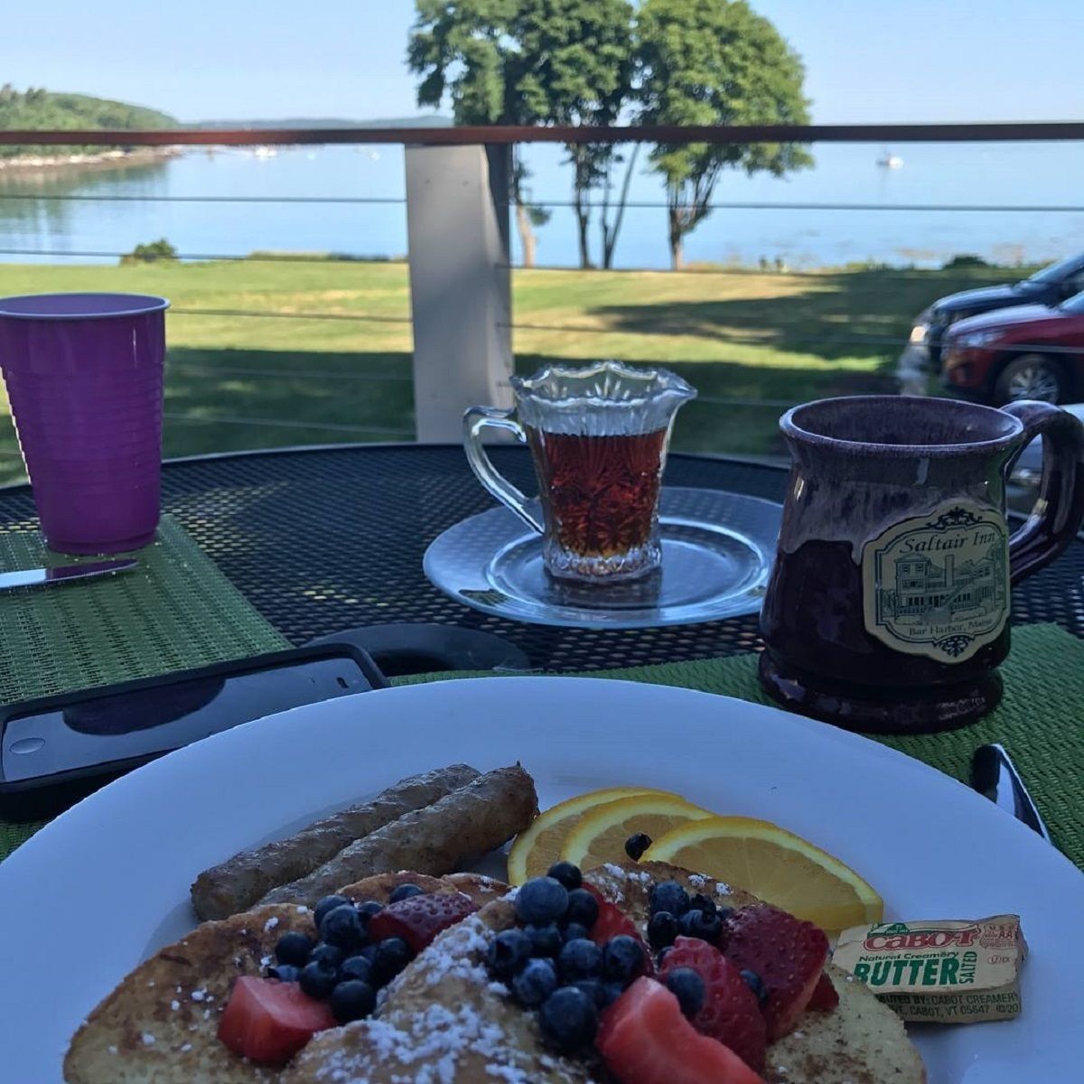 A breakfast setting with pancakes, blueberries, strawberries, and orange slices on a plate, a pitcher of tea, a mug, and a bottle of butter.