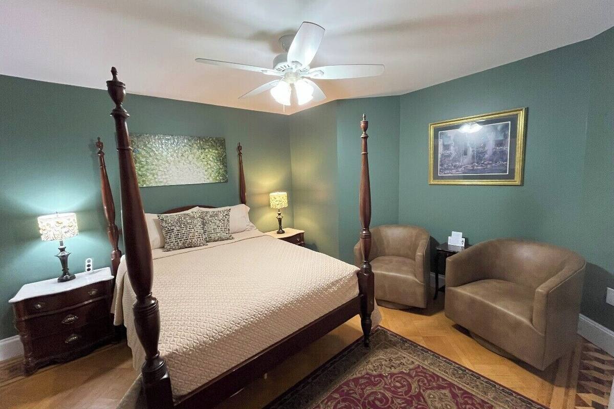 A bedroom with a four-poster bed, a ceiling fan, a nightstand with a lamp, a chair, and a sofa.