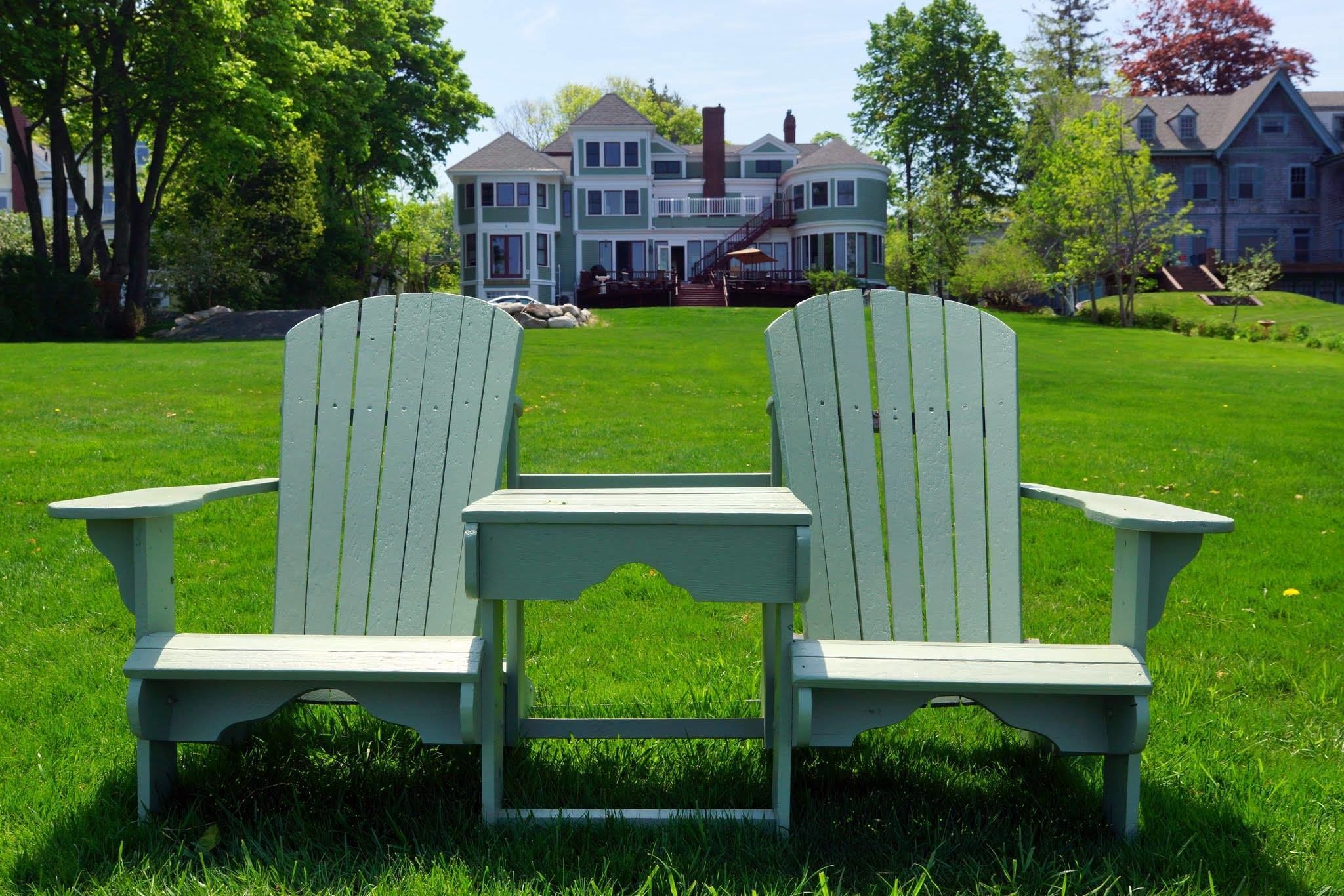 Two Adirondack chairs facing a large, two-story house with a front porch and a staircase leading to the second floor.