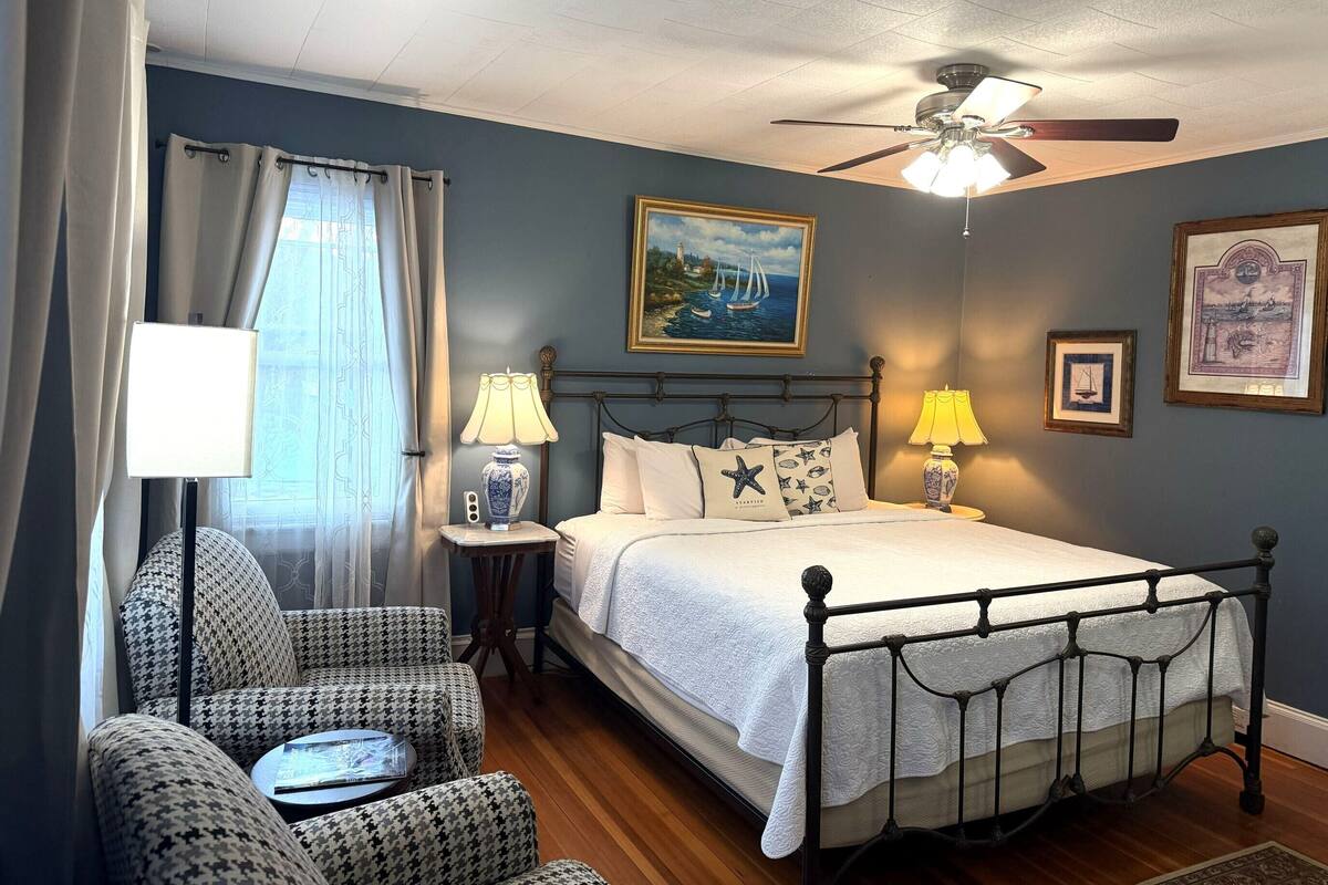A neatly arranged bedroom with a bed, two bedside tables with lamps, a chair, a ceiling fan, and a painting on the wall.
