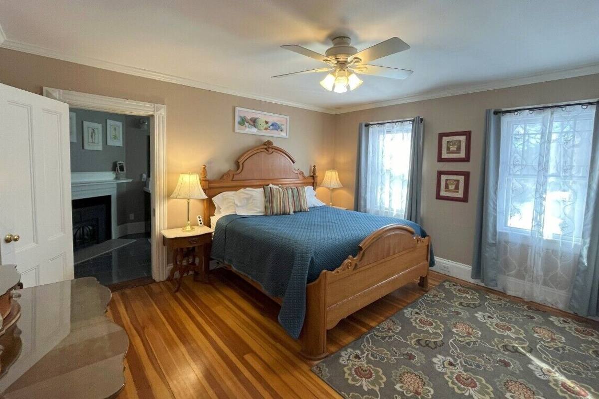 A bedroom with a wooden bed, a ceiling fan, a fireplace, and a view of the outdoors through a window with curtains.