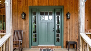 Un porche de madera con una puerta verde, dos mecedoras y una pequeña mesa.