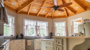 Una cocina con vigas de madera en el techo, ventilador de techo y ventanas grandes con cortinas.