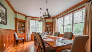 Un comedor con una mesa de madera, sillas y una araña. Se ven ventanas con cortinas y vistas a árboles desde el exterior.