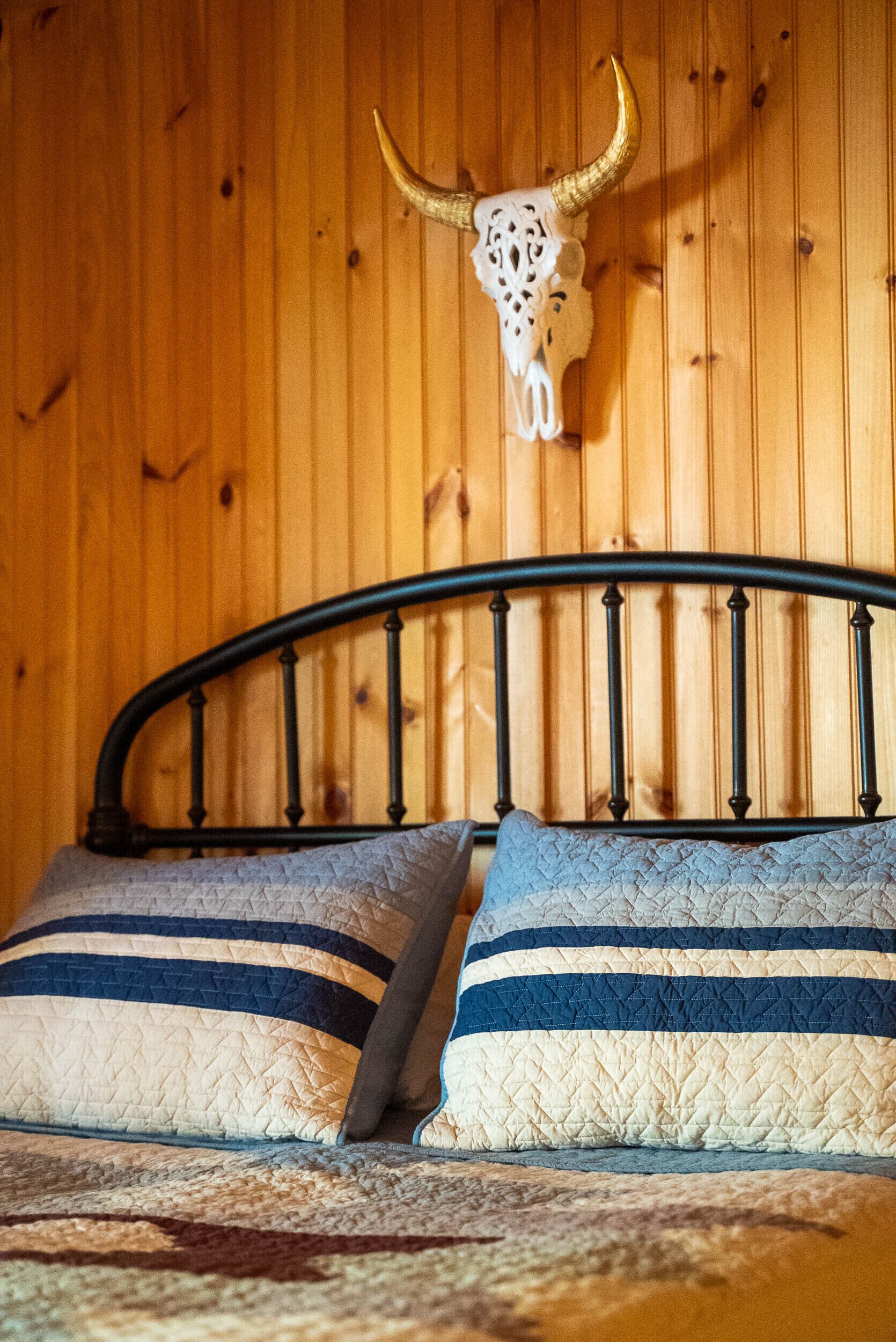 Ein Bett mit Metallkopfteil, zwei Kissen mit Streifenmuster und ein Kuhschädel, der an der Wand befestigt ist.
