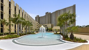 A swimming pool with a central fountain, surrounded by palm trees and a modern building complex.