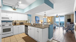 A modern kitchen with white cabinets, a granite countertop, and a built-in microwave. There is a dining area with a table and chairs, and a view of the ocean through large windows.