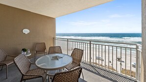 A balcony with wicker chairs and a round table, overlooking a beach with waves and a clear sky.