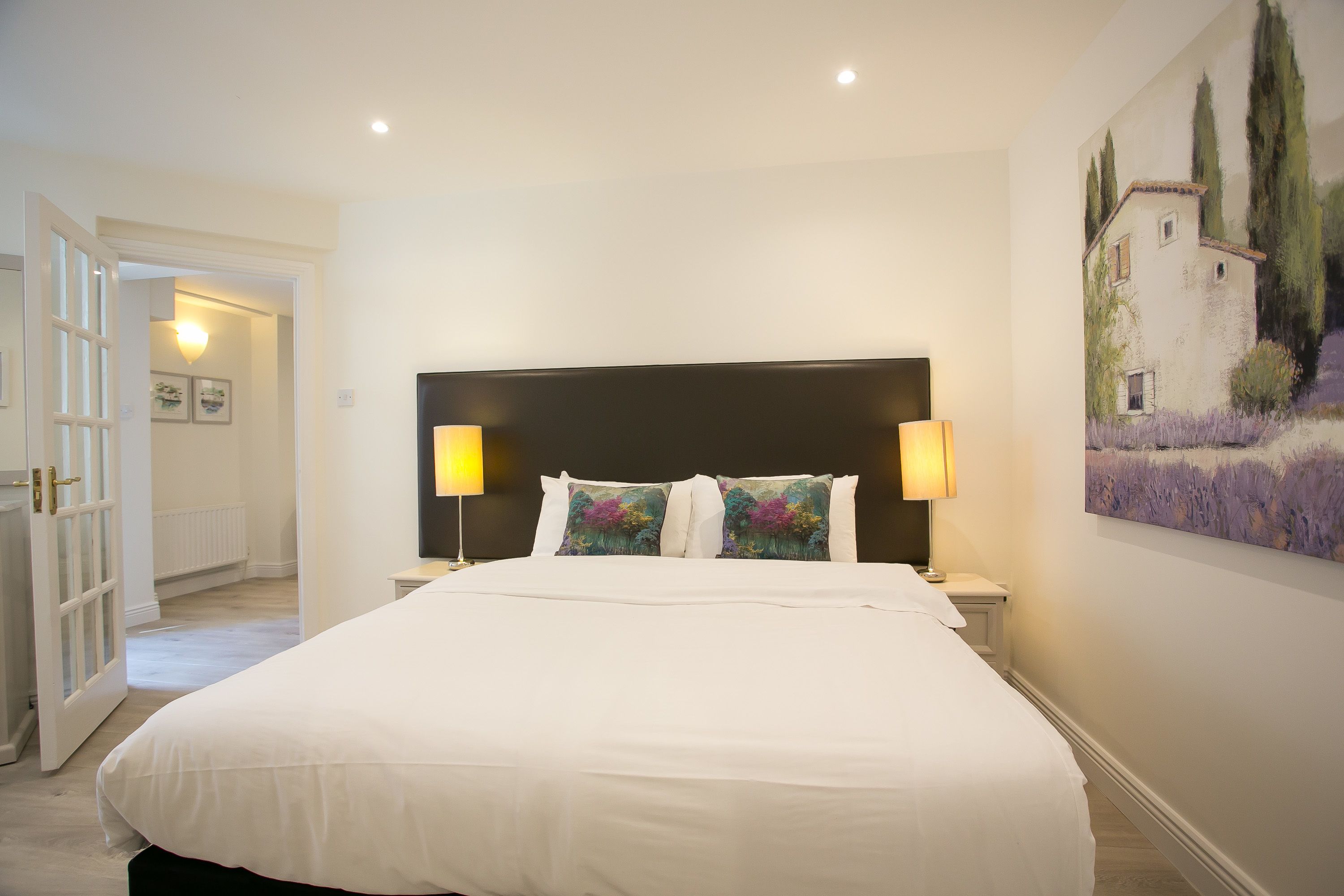 Una cama bien hecha con ropa blanca, dos lámparas de noche y un cuadro de una casa en un campo en la pared.