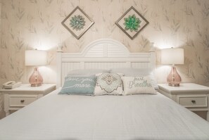 A neatly made bed with decorative pillows, two bedside lamps, and a floral wallpaper pattern. - Scenic Ocean front first floor pool view spacius condo for up to 4 adults. (Garden City)