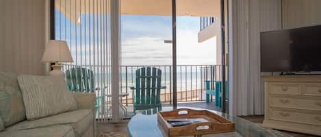 Habitación de hotel con sofá, una mesita de vidrio, televisor, balcón con vistas a la playa y una mesita auxiliar de madera.
