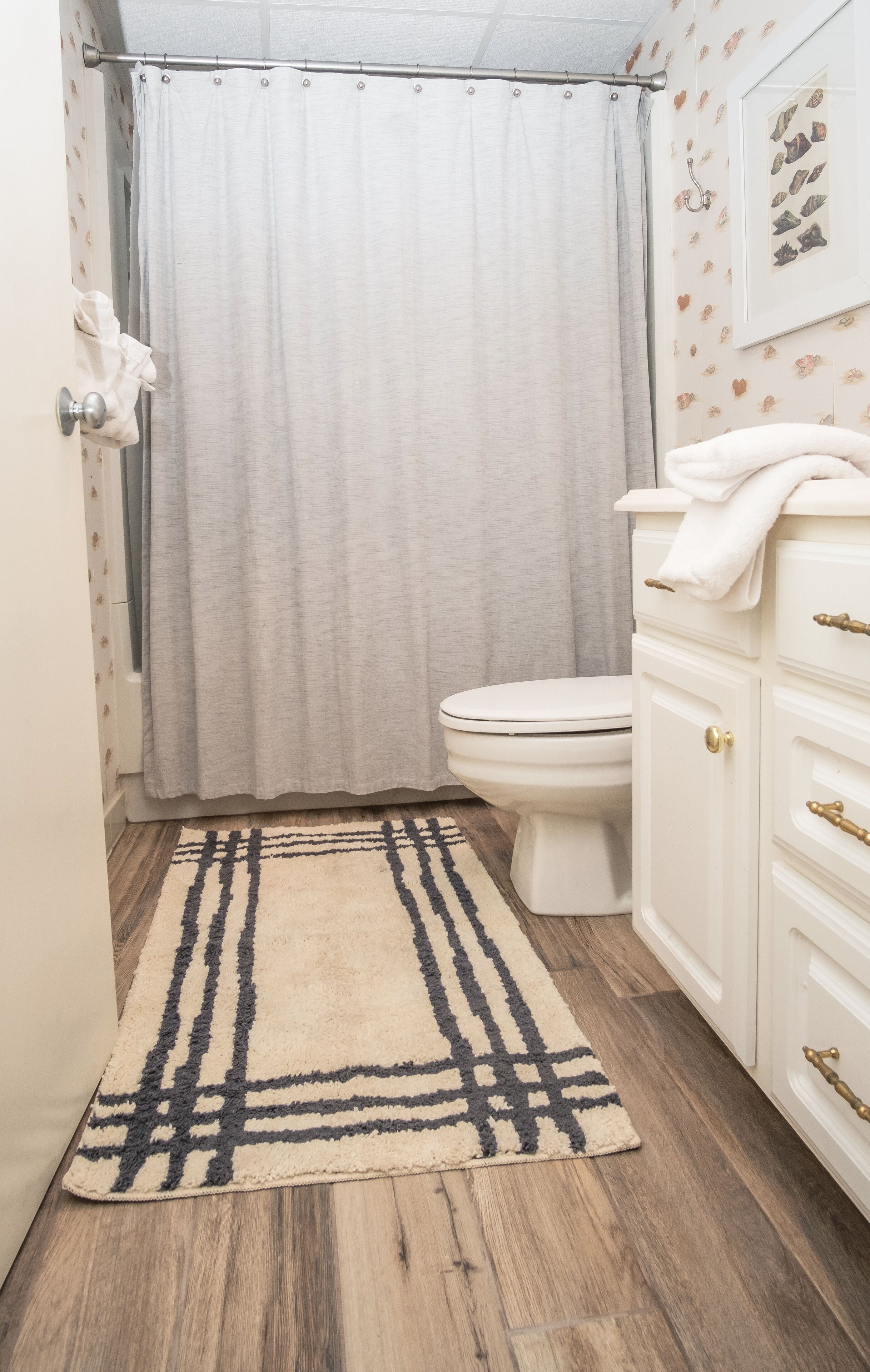A bathroom with a toilet, a shower curtain, a patterned rug, and a white cabinet with gold handles.