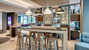 A modern bar area with a central island, bar stools, pendant lighting, and a bookshelf with decorative items. - Premier Inn Frankfurt Westend (Frankfurt)