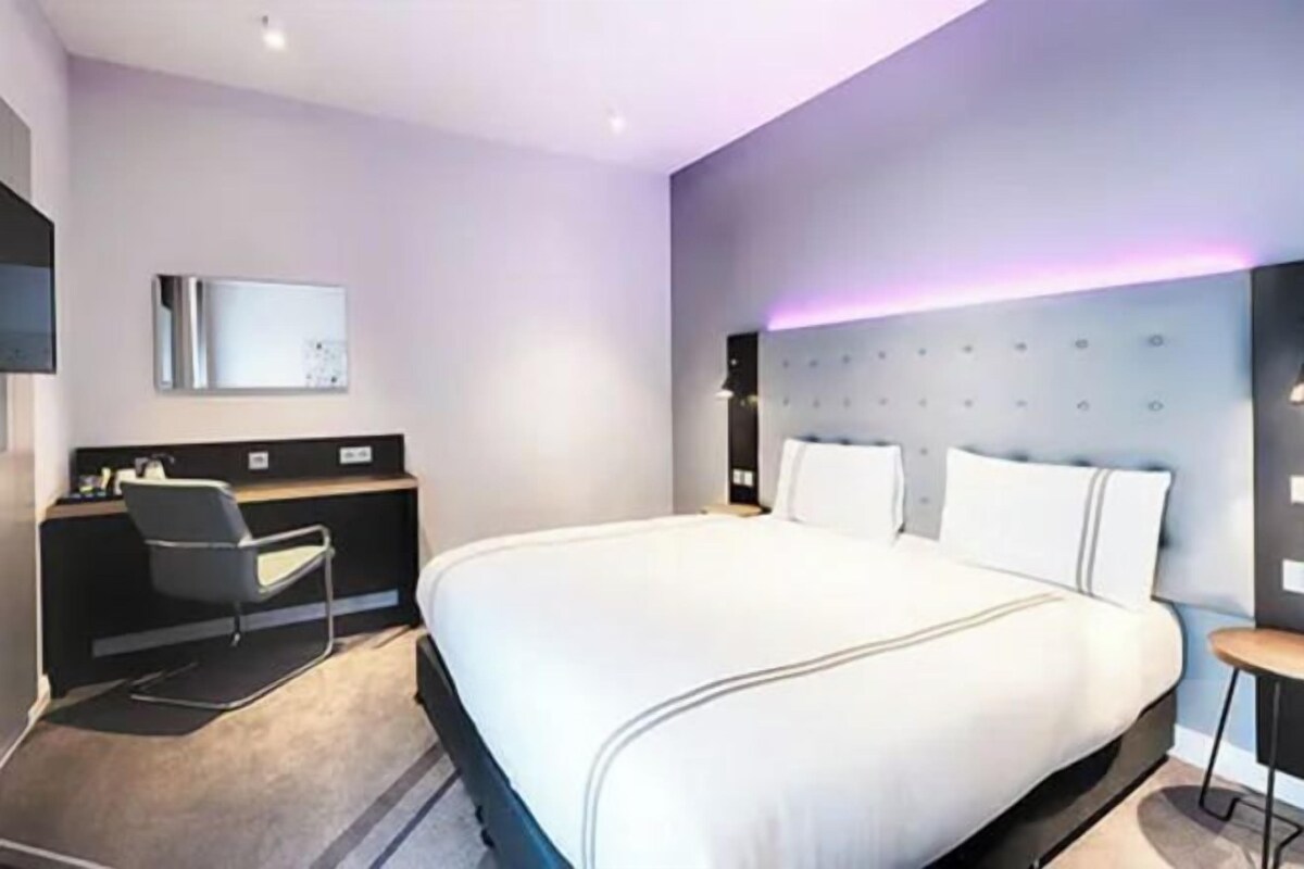 Una habitación de hotel moderna con una cama grande, un escritorio con silla, un televisor de pantalla plana y un cabecero con iluminación ambiental.