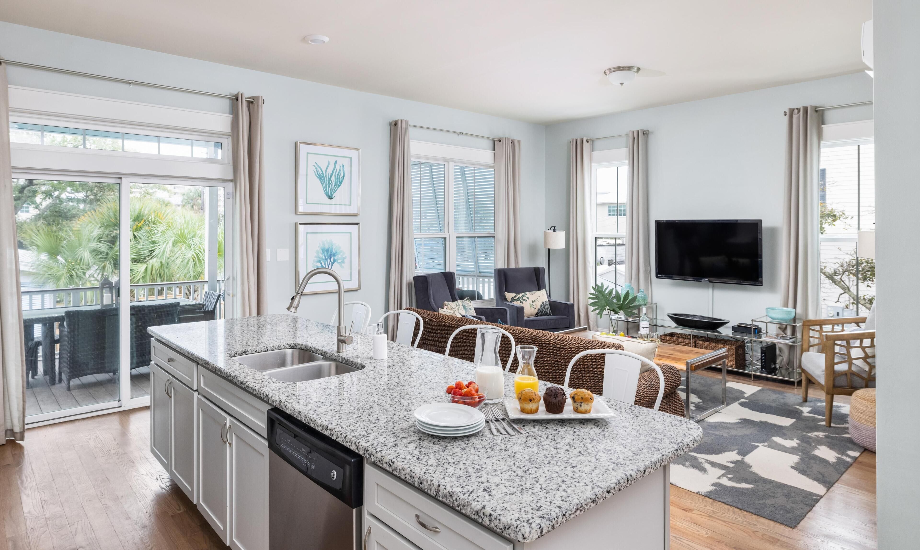 A modern kitchen with a granite countertop, a dishwasher, and a dining area with a table and chairs. There is a TV mounted on the wall and a balcony with a view of greenery.