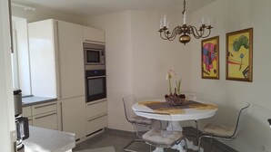 Een compacte keuken met een magnetron, oven en een eettafel met stoelen. Er hangen twee kunstwerken in lijsten aan de muur.
