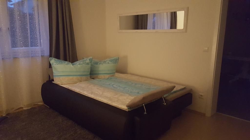 A small bedroom with a bed, a window with curtains, a mirror above the bed, and a door to the right.