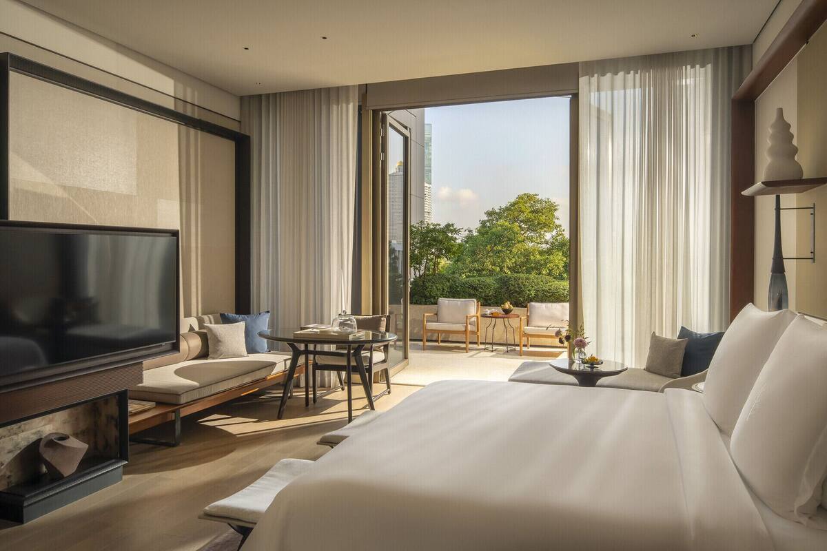 A modern hotel room with a large bed, a TV, a seating area with a table, and a balcony with a view of the city.