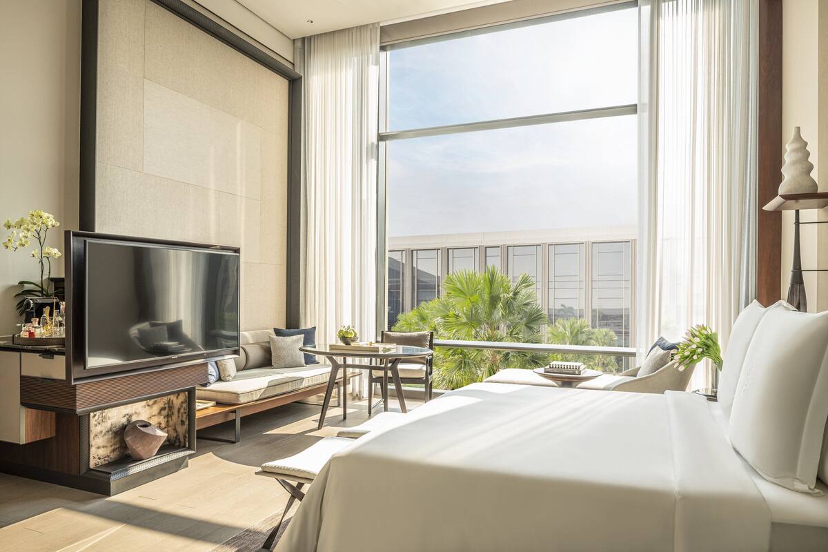 A modern hotel room with a large bed, a flat-screen TV, a balcony with a view of palm trees, and a seating area with a small table and chairs.