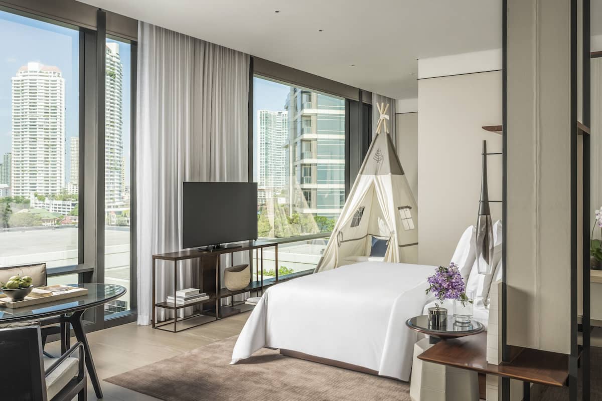 A modern hotel room with a large bed, a TV, a balcony with a view of tall buildings, and a small table with a vase of flowers.