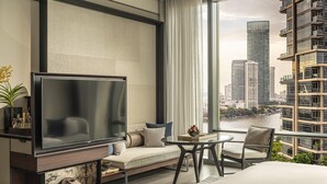 A modern hotel room with a large TV, a sofa, a dining table, and a view of the cityscape and river. - Four Seasons Hotel Bangkok at Chao Phraya River (Bangkok)