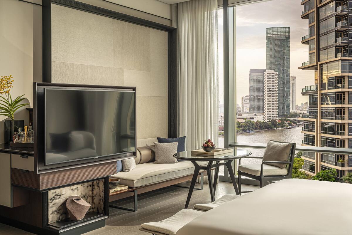 A modern hotel room with a large TV, a sofa, a dining table, and a view of the cityscape and river.