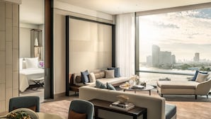 A modern hotel room with a large window offering a city view, a comfortable seating area, and a dining table. - Four Seasons Hotel Bangkok at Chao Phraya River (Bangkok)