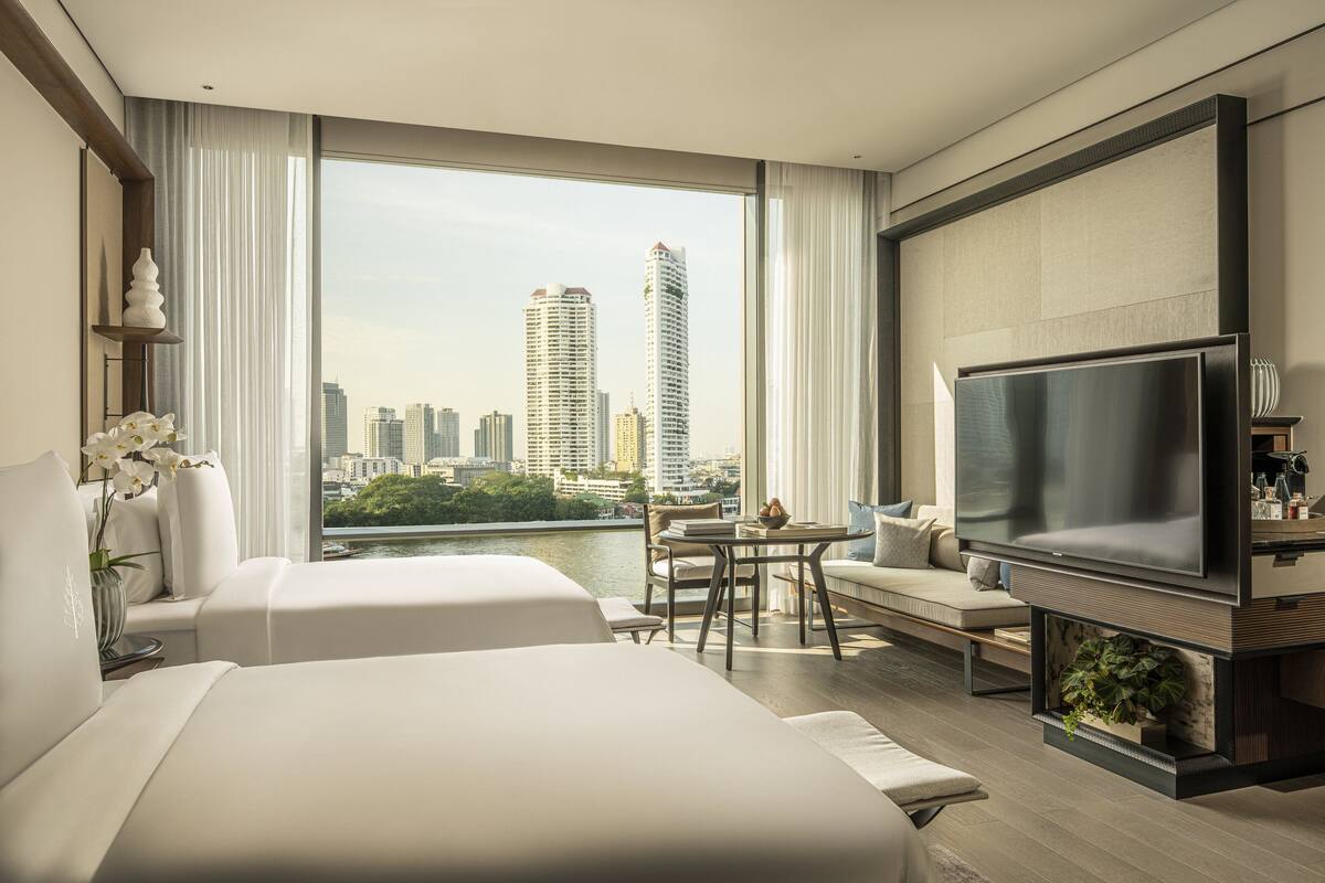 A modern hotel room with two beds, a TV, a dining area, and a view of the city skyline.