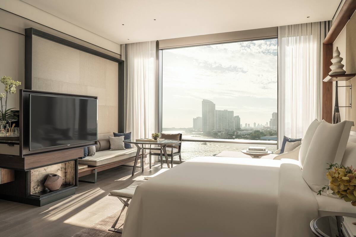 A modern hotel room with a large bed, a flat-screen TV, a seating area with a table, and a view of the city skyline.