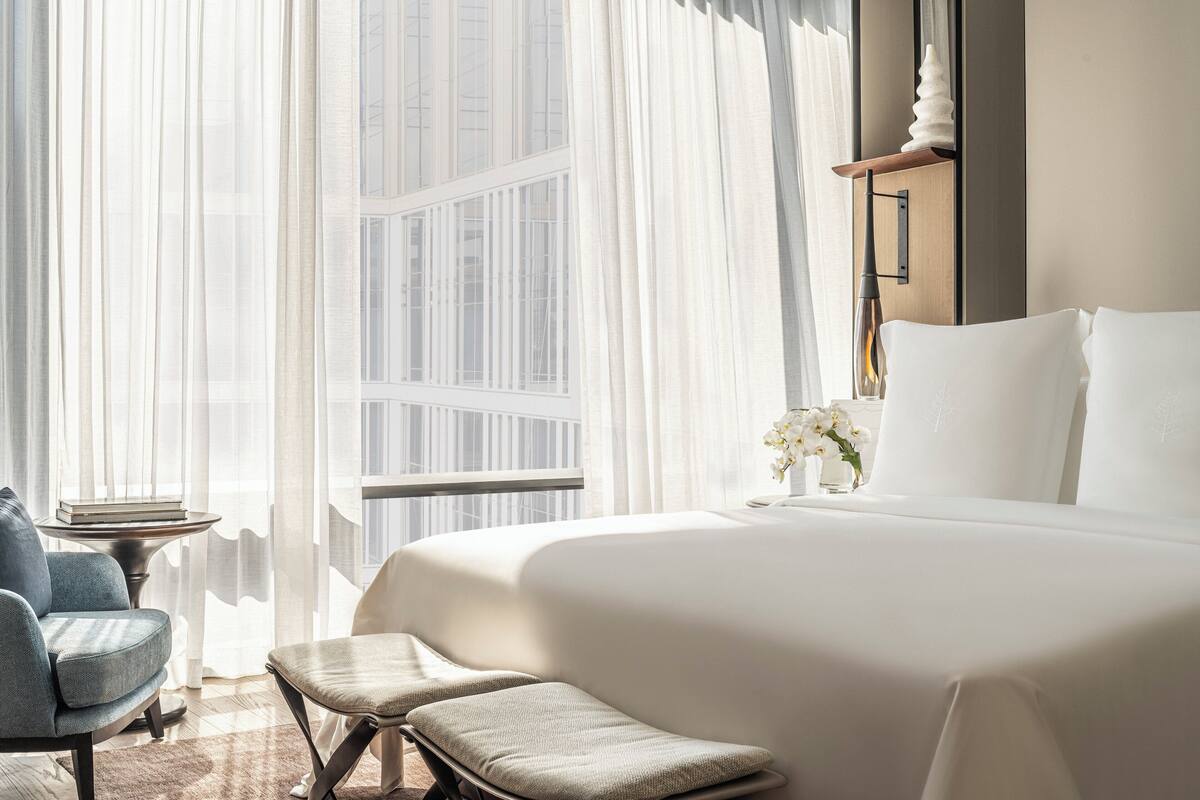 A hotel room with a large bed, a sitting area with a chair and a small table, and a view of the city through sheer curtains.