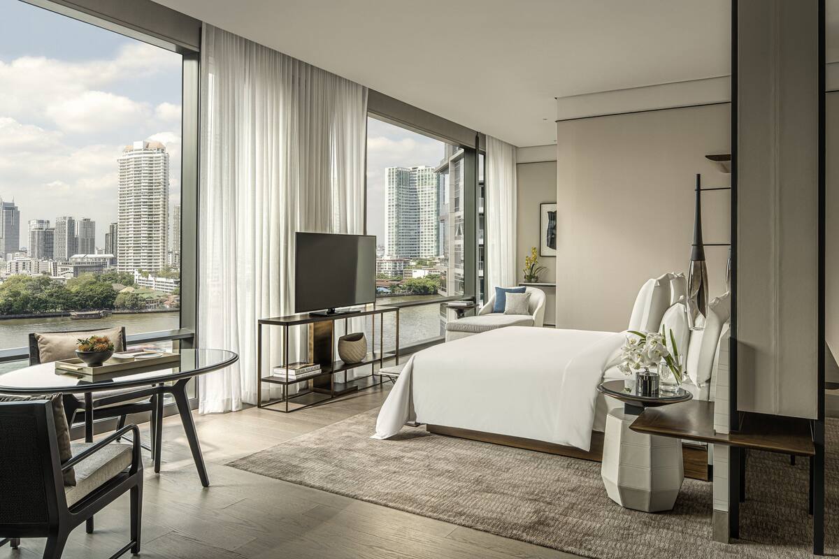 A modern hotel room with a large bed, a TV, a dining area, and a view of the city skyline.
