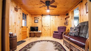 Un interior de cabaña acogedor con paredes de madera, un ventilador de techo, un televisor, un sillón morado, un sofá de cuero marrón y una alfombra estampada.