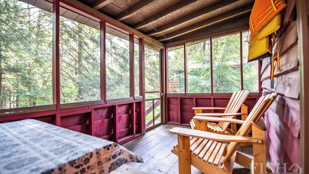 Una cabaña de madera con un porche, sillas de madera, una mesa y ventanas amplias que dan a un bosque.