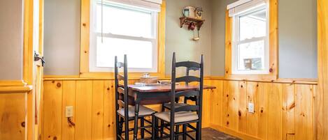 A dining area with a wooden table and chairs, two windows with blinds, and a shelf with decorative items.
