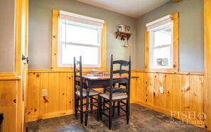 Un coin repas avec une table et des chaises en bois, deux fenêtres équipées de stores et une étagère ornée d’objets décoratifs.