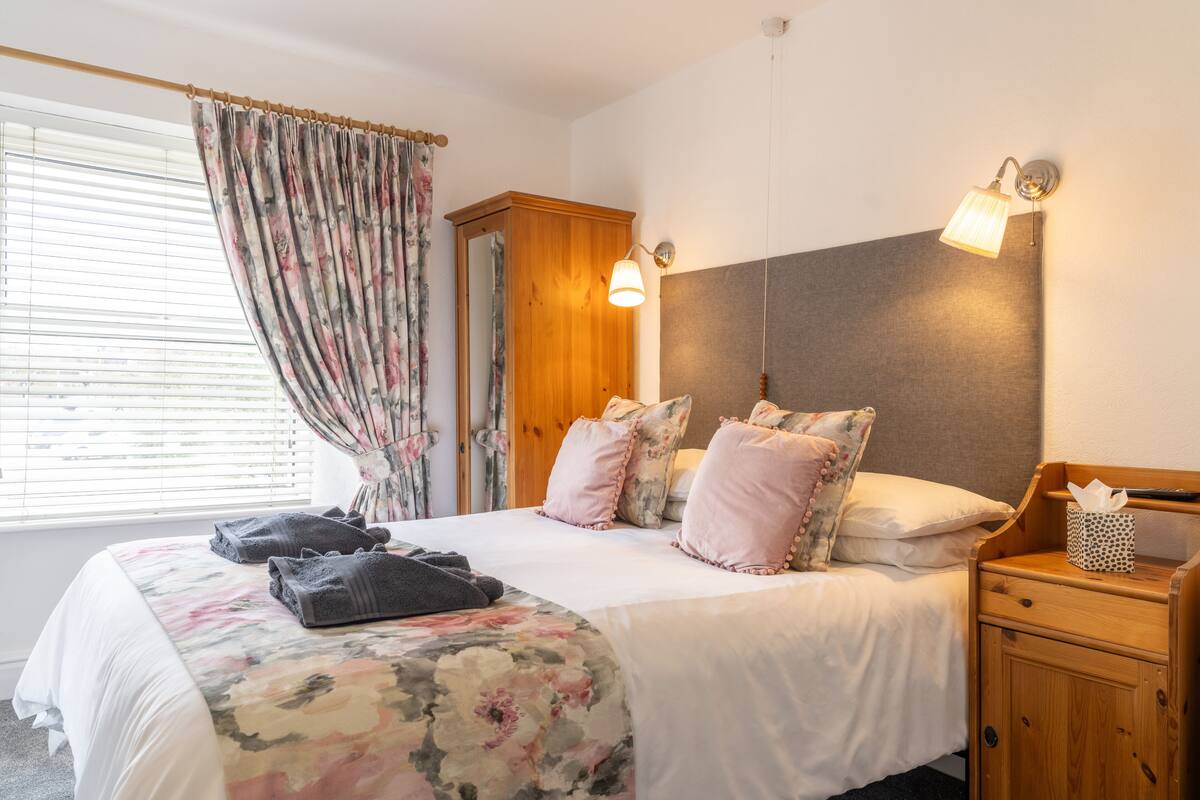 A bedroom with a bed, pillows, a nightstand, a mirror, and a window with curtains.