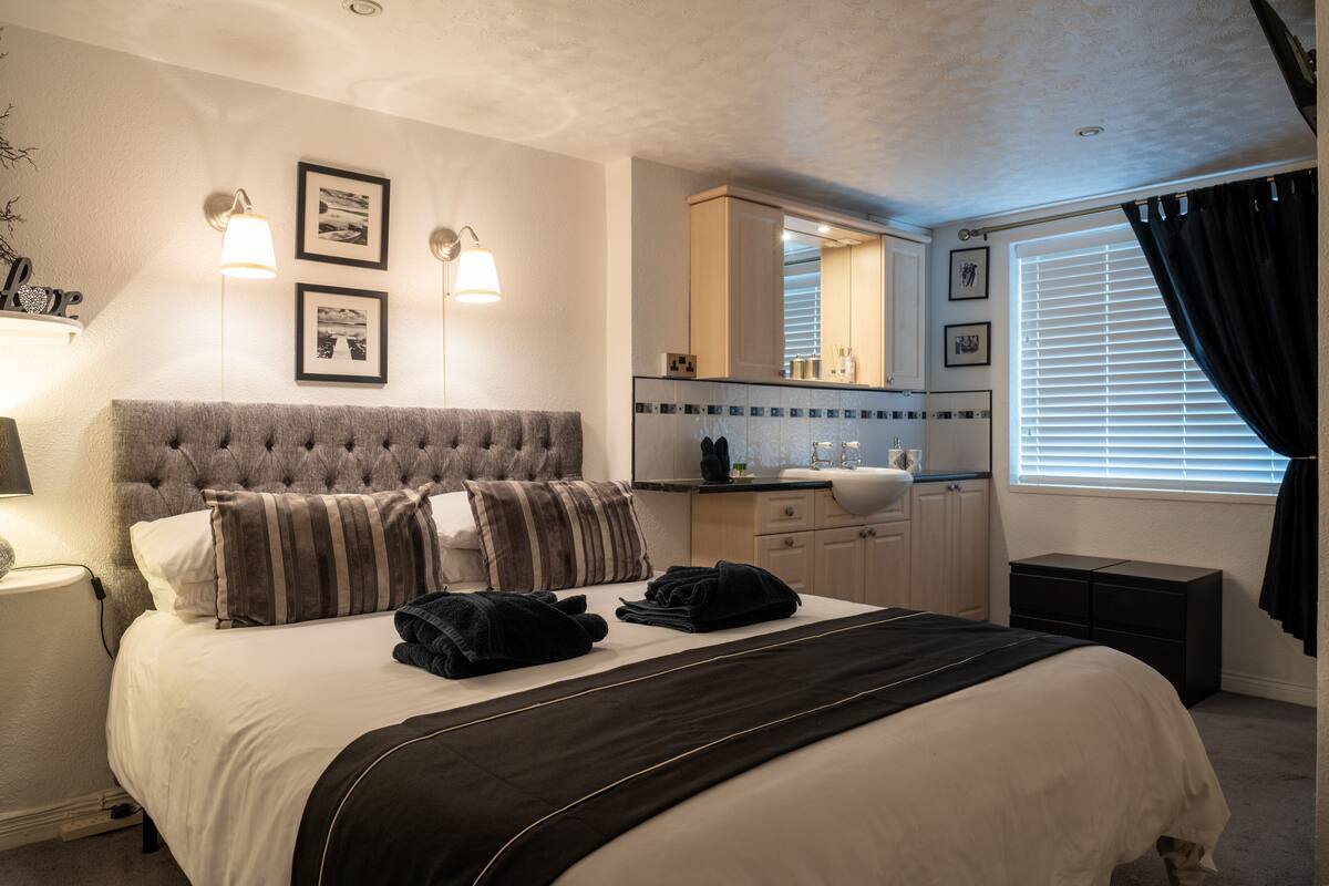 A hotel room with a bed, a vanity with a mirror, a window with curtains, and wall-mounted lights.