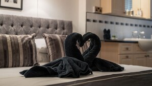 A neatly made bed with a towel arranged in a heart shape, a tufted headboard, a framed picture on the wall, and a kitchen area in the background. - Belmont House Beer - Adults Only (Seaton)