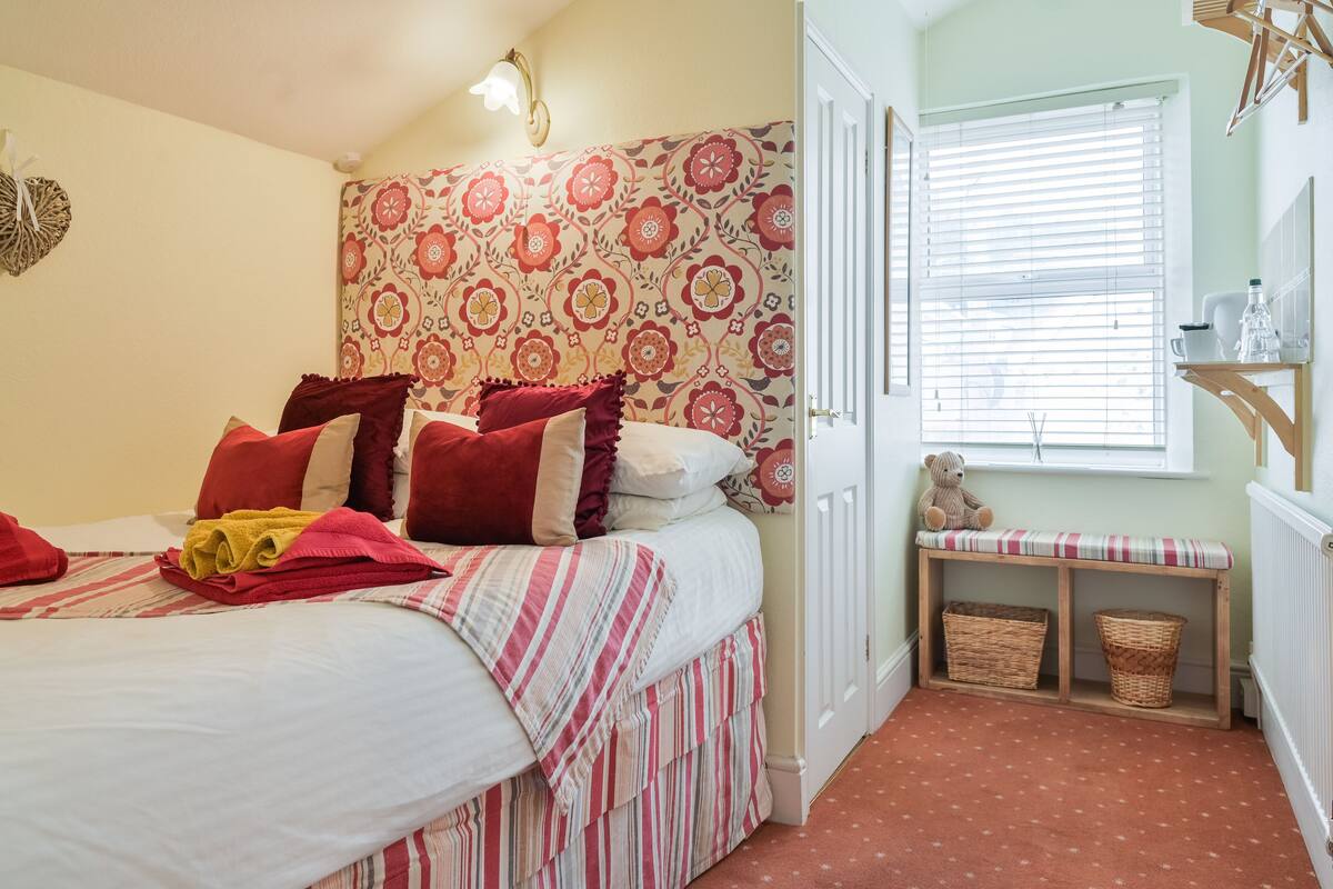 A bedroom with a bed, pillows, a bedside table, a window, and a wall with a floral pattern.