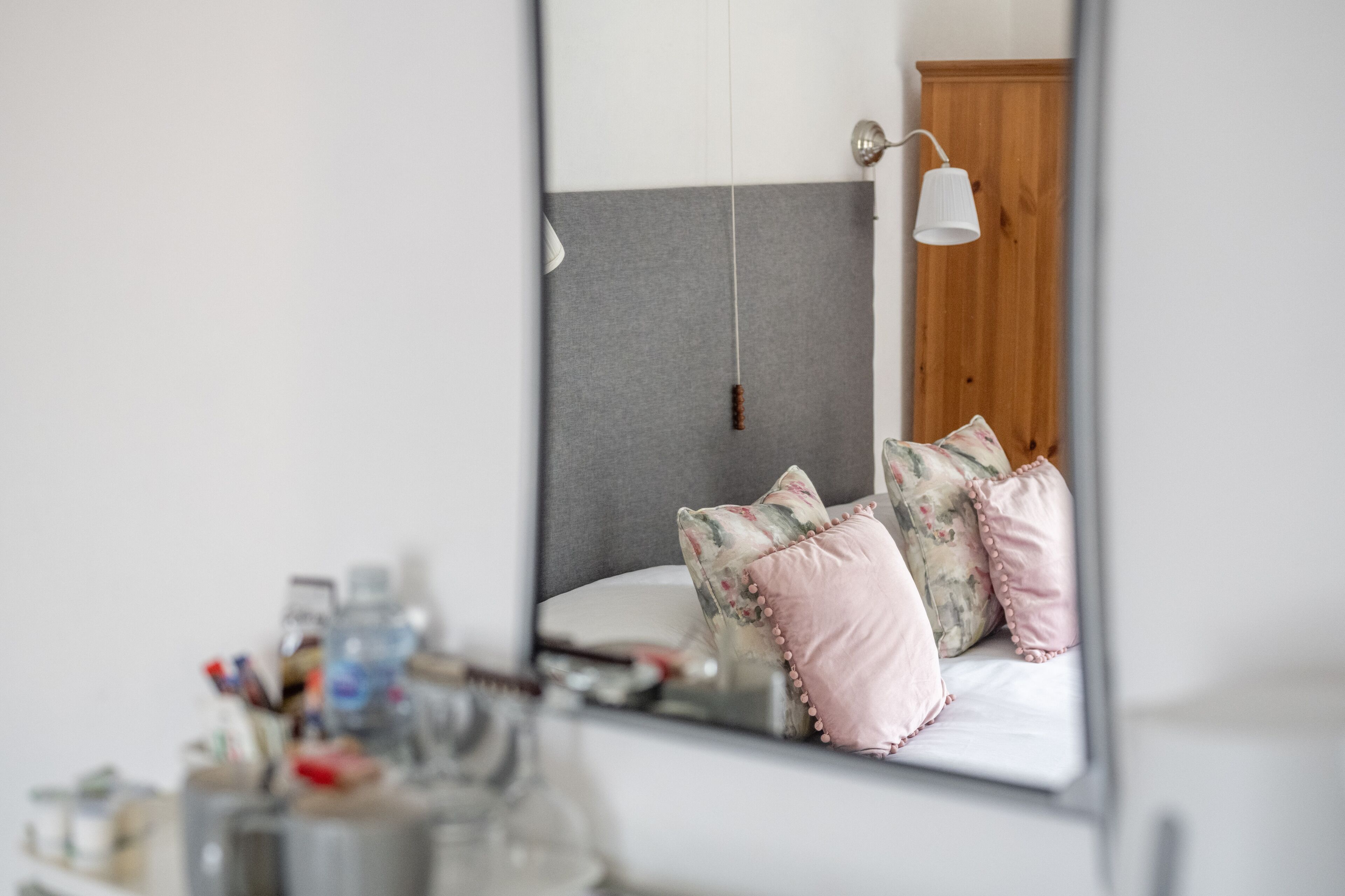 A bedroom with a large mirror, a bed with floral and pink pillows, a wooden door, and a bedside table with toiletries.