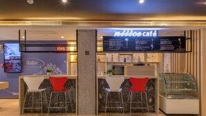 A modern cafe interior with a counter, two red and two white bar stools, a menu display, and a glass display case. - Ibis Wuhan Hubu Alley Hotel (Wuhan)