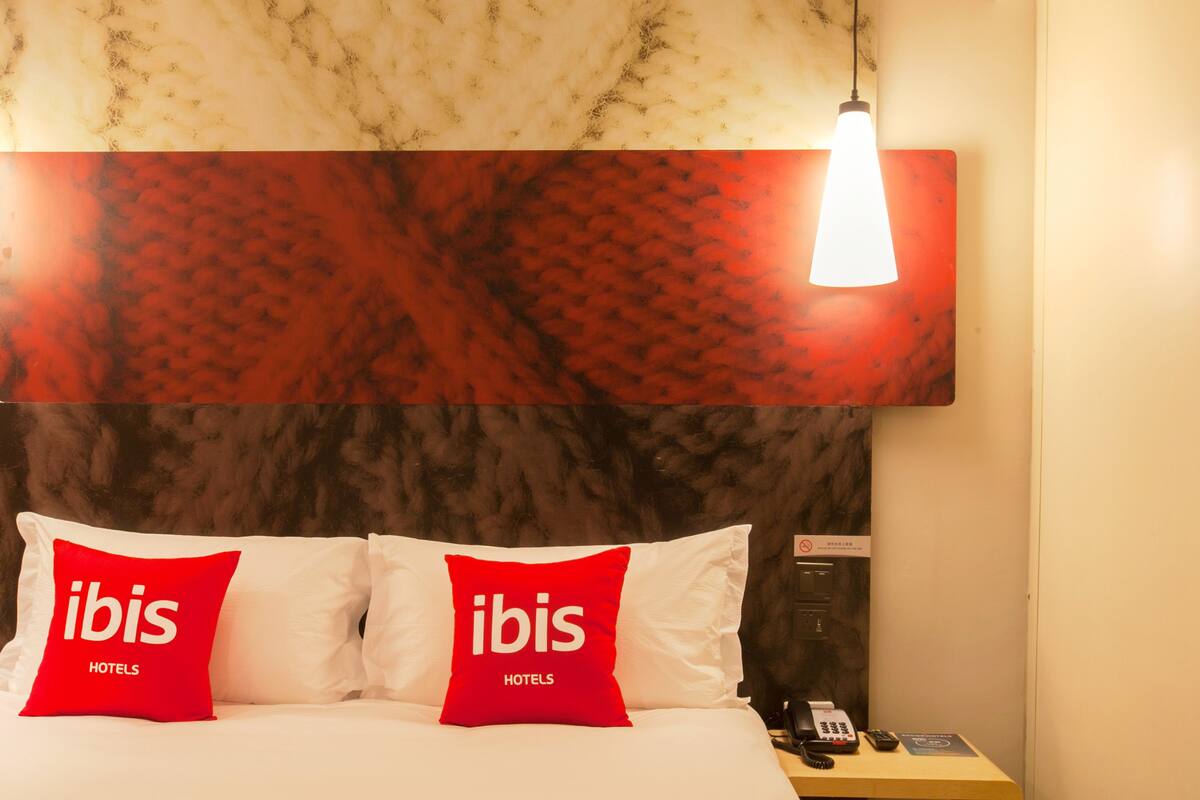 A hotel room with a marble headboard, two red pillows with "ibis HOTELS" text, a bedside telephone, and a wall-mounted light fixture.