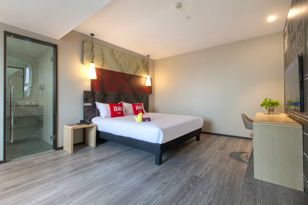 A modern hotel room with a large bed, a small bedside table, a desk, and a glass door leading to a shower area.